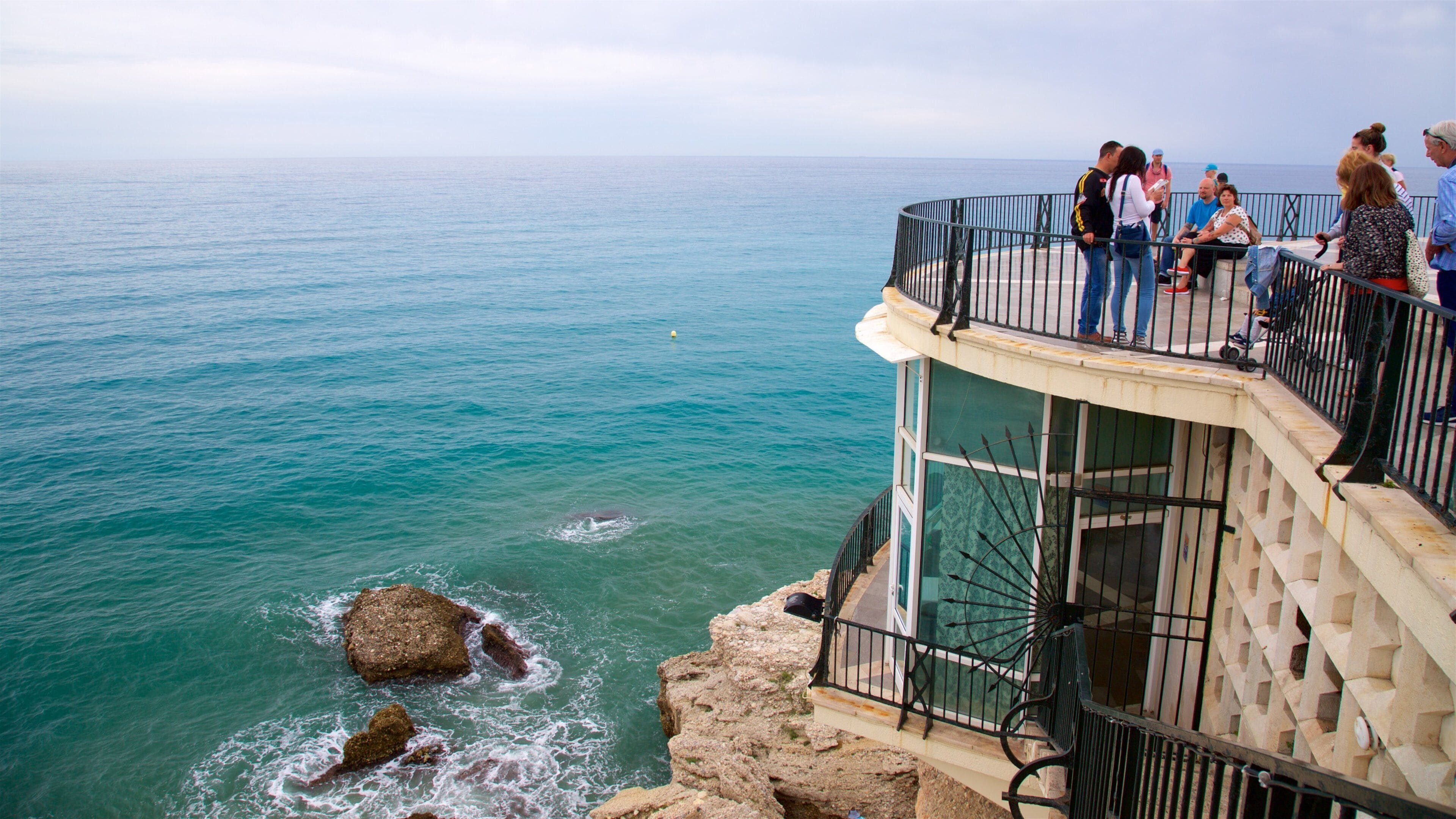 Ein Küstenblick mit Menschen, die aufs Meer schauen, einem Felsen und einem gläsernen Gebäude.