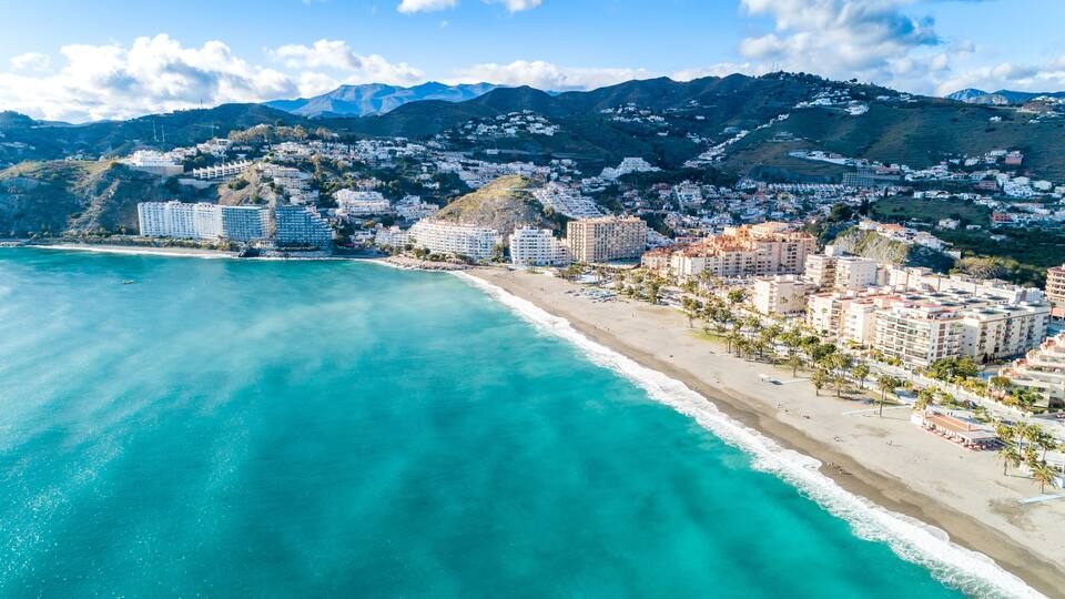 A coastal city with a beach, buildings, and mountains in the background.