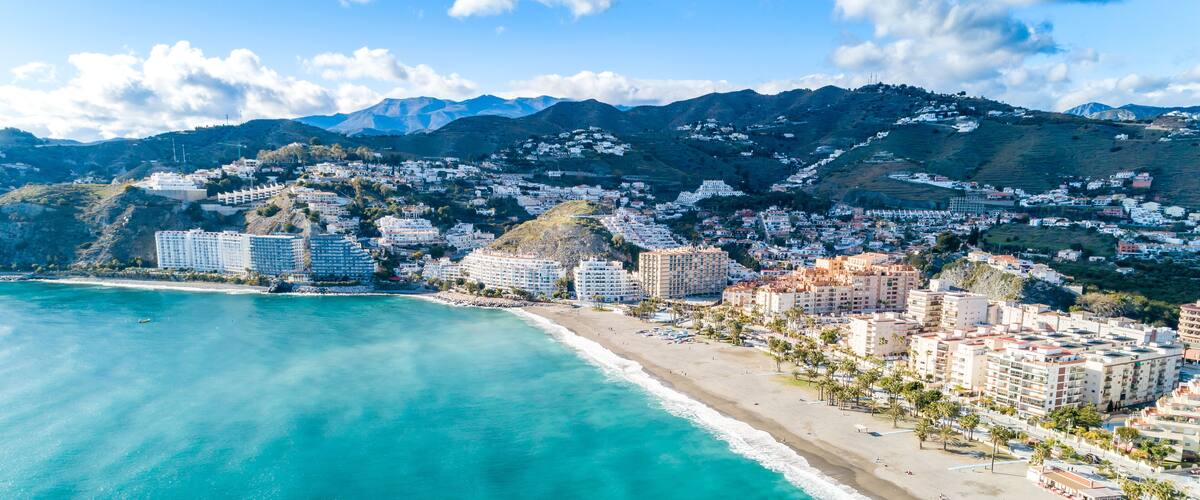 A coastal city with a beach, buildings, and mountains in the background.