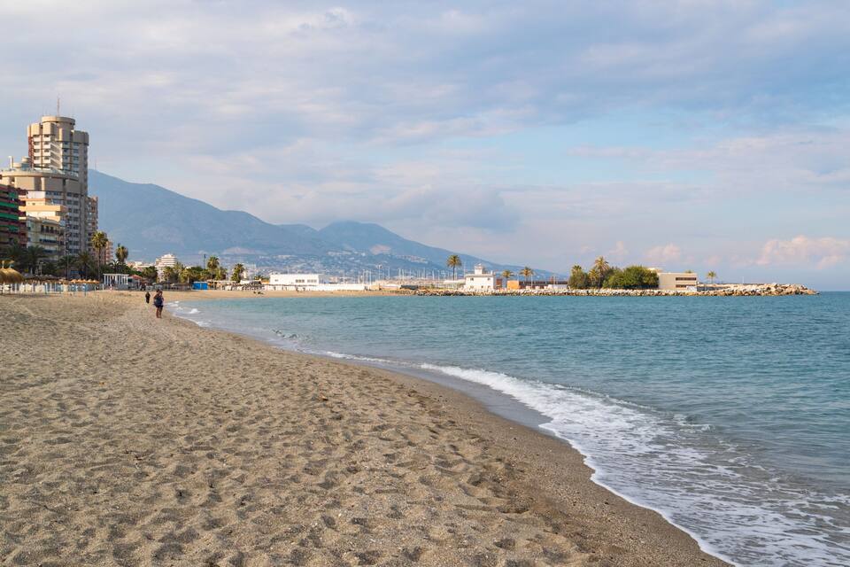 A beach with a rocky breakwater, palm trees, and a mountain in the background.