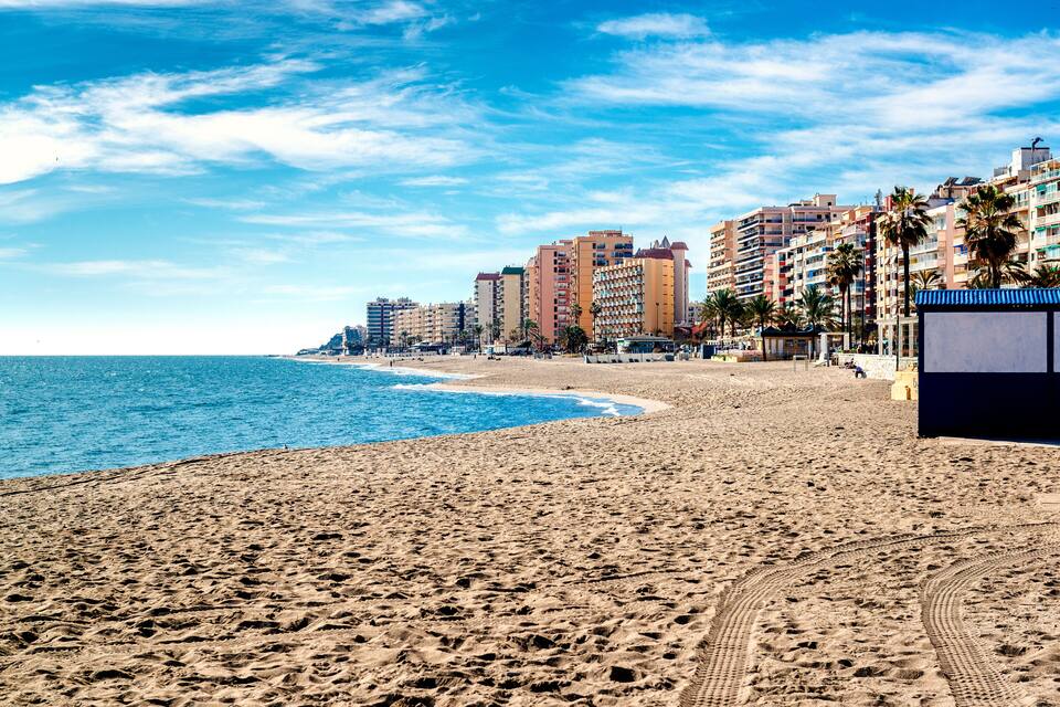 A sandy beach with footprints, a clear blue sky, and tall buildings in the background.