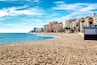 A sandy beach with footprints, a clear blue sky, and tall buildings in the background.