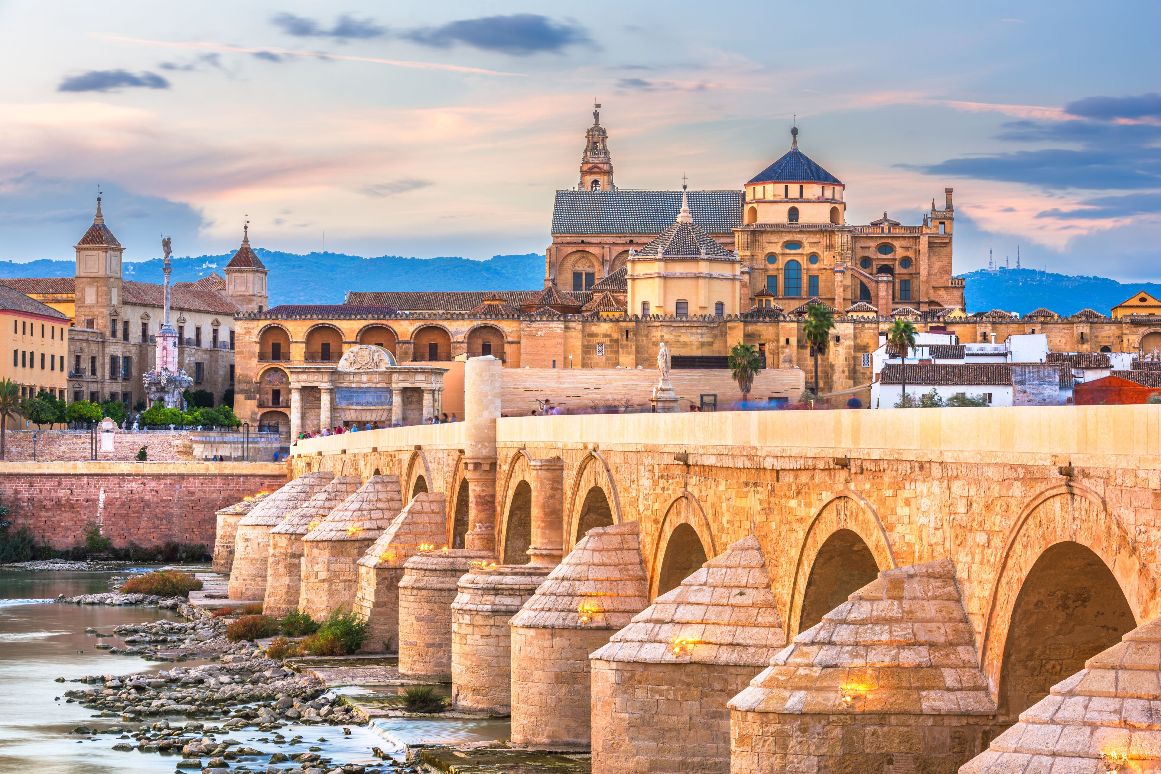Un puente de piedra con arcos sobre un río, un edificio histórico con una torre de campanas y un paisaje montañoso al fondo.