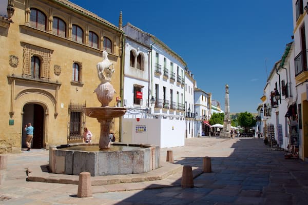 Uma praça histórica com uma fonte, ruas de paralelepípedos e arquitetura europeia tradicional.
