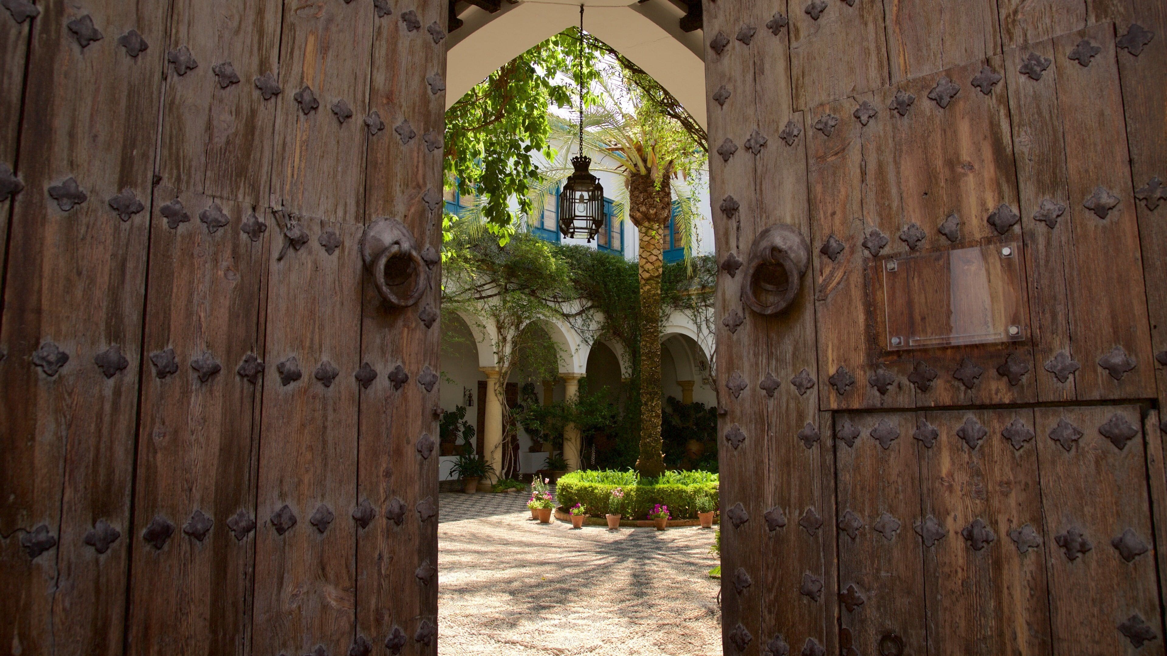 A wooden gate with metal studs, leading to a courtyard with arches, a lantern, and potted plants.