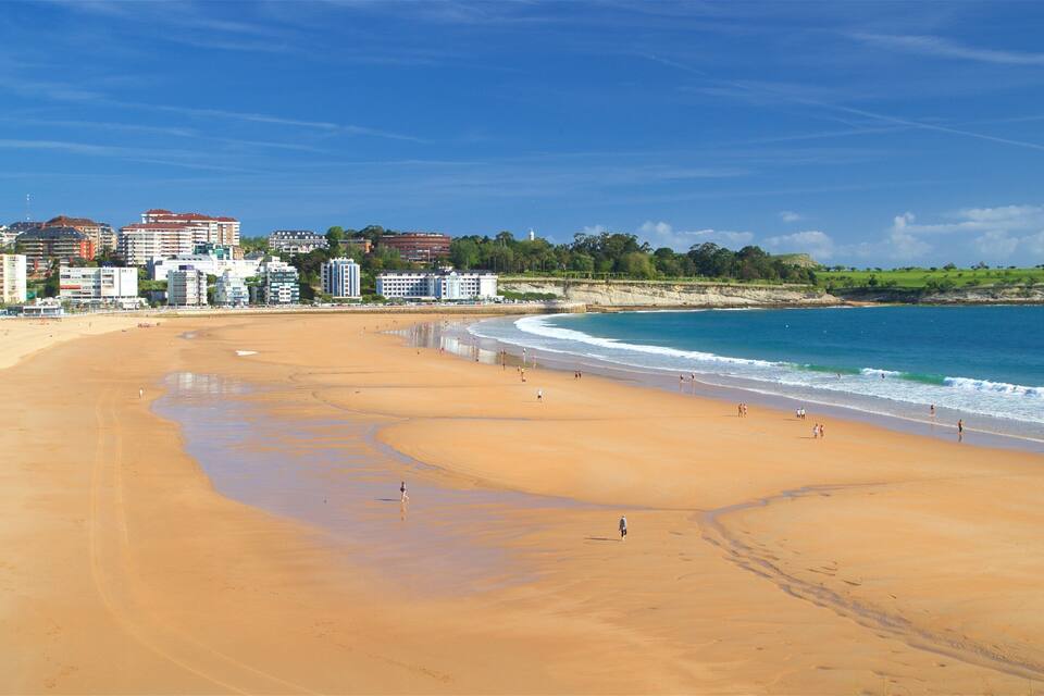 A wide sandy beach with a clear blue ocean, a lighthouse, and a cityscape in the background.