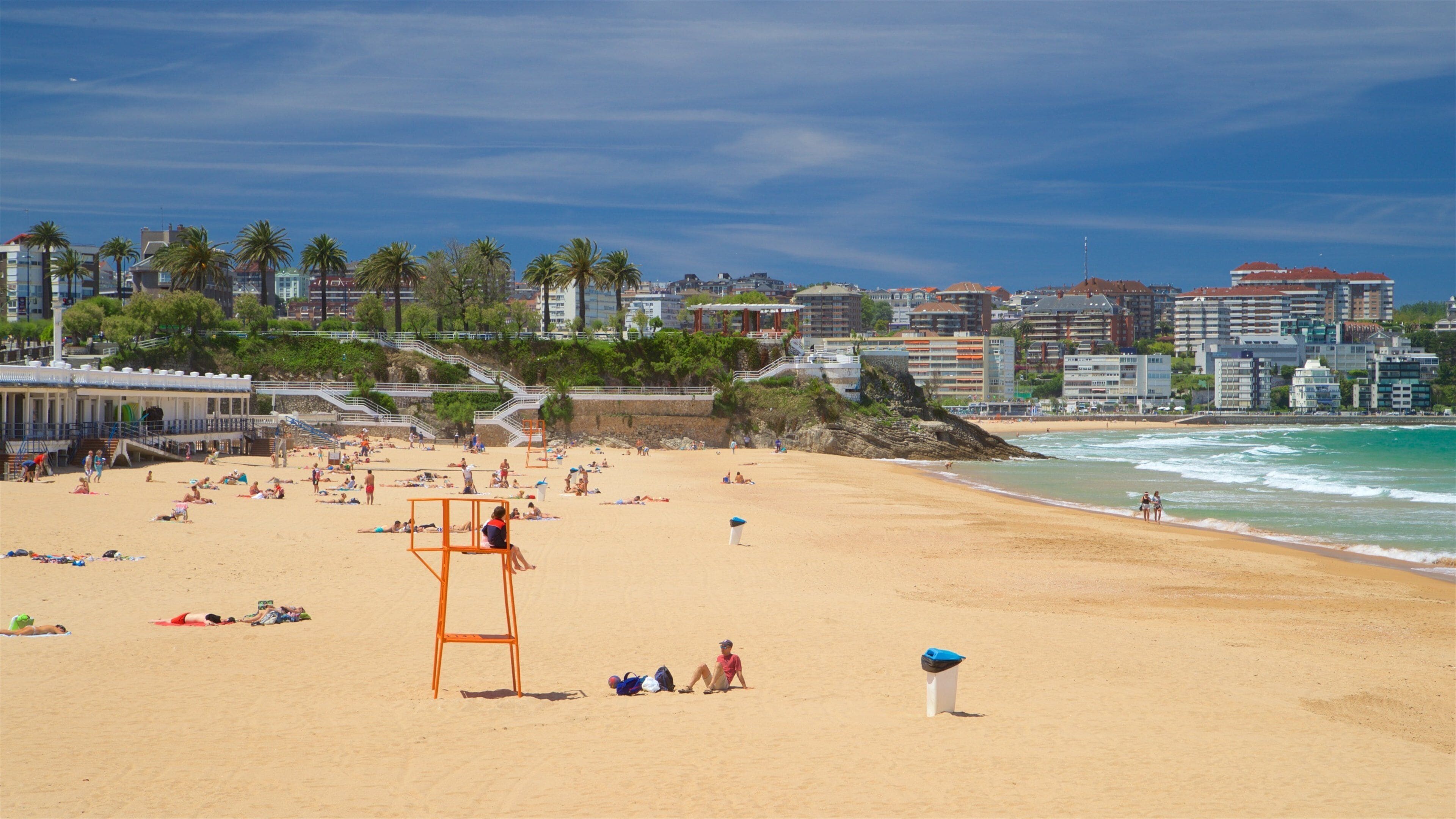 Une plage où l’on voit des gens se prélasser au soleil et nager, des palmiers et des bâtiments en arrière-plan.