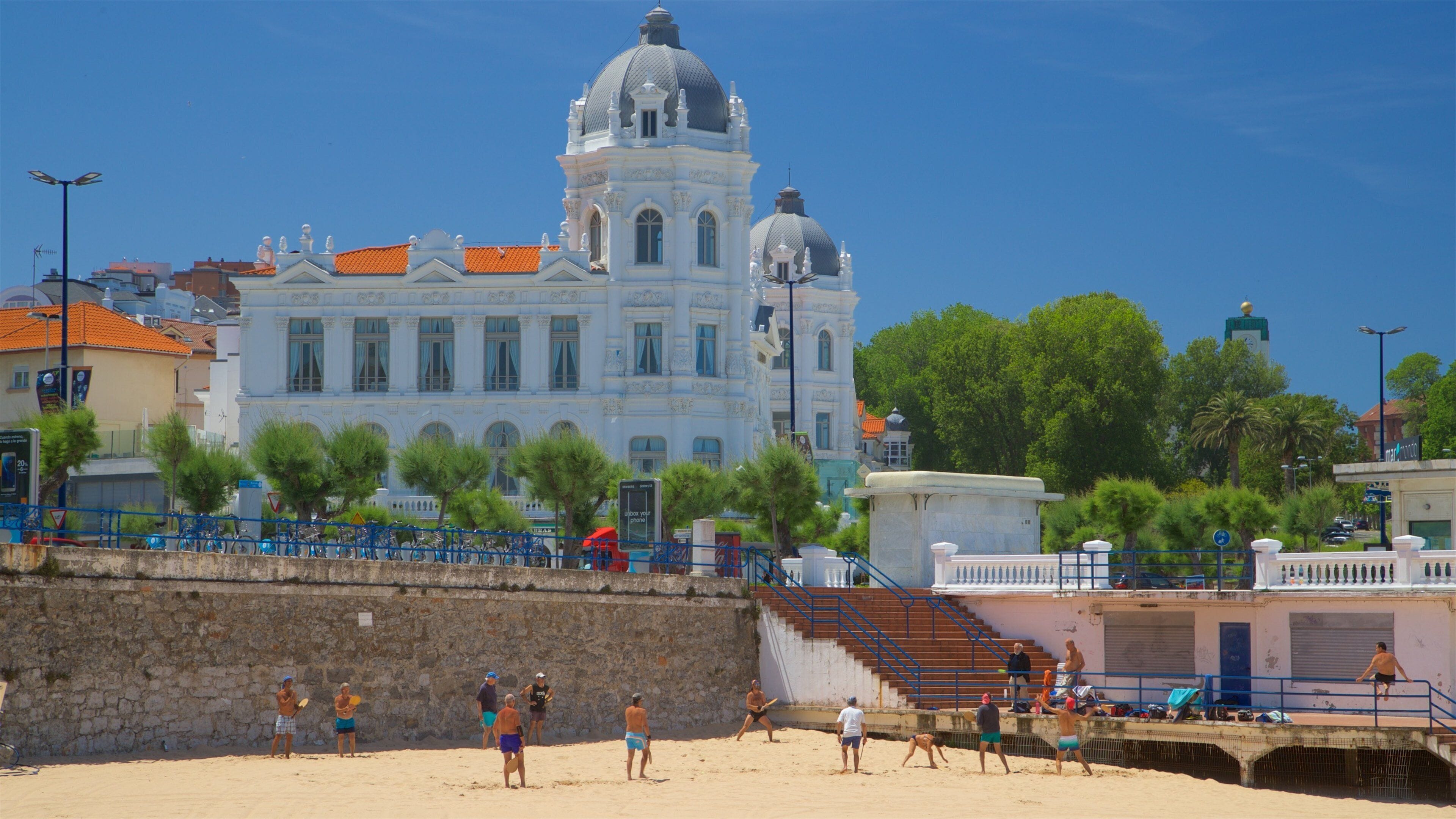 Una playa con gente jugando al voleibol, un muro de piedra y un gran edificio blanco con una cúpula al fondo.