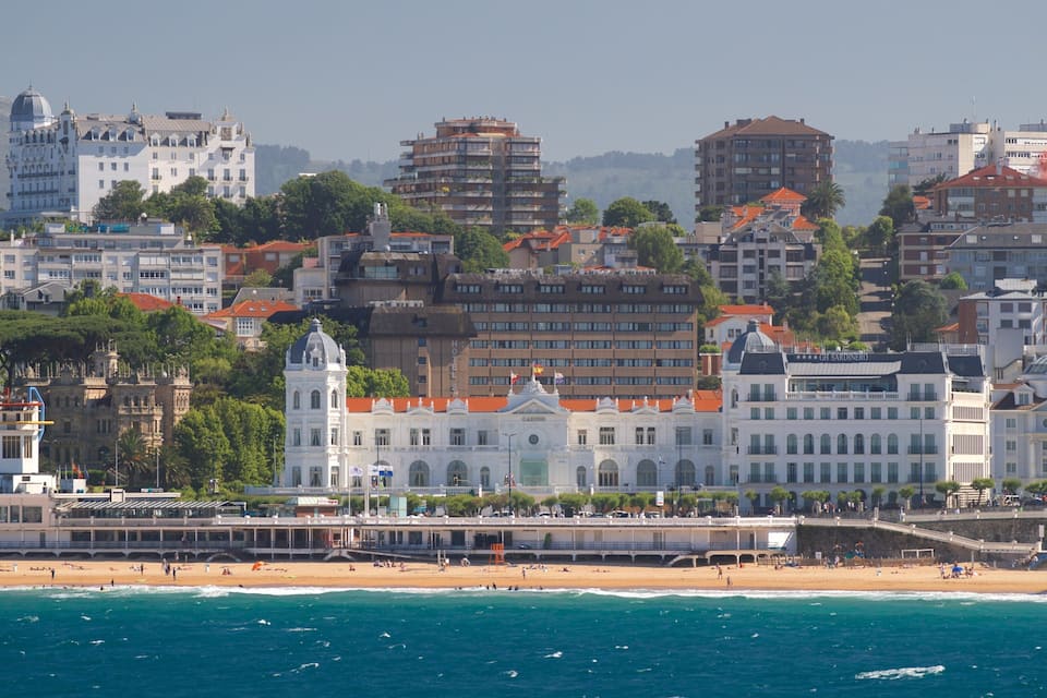 Un paysage urbain côtier avec un imposant bâtiment blanc surmonté d’un dôme, une plage et une vue sur la mer.