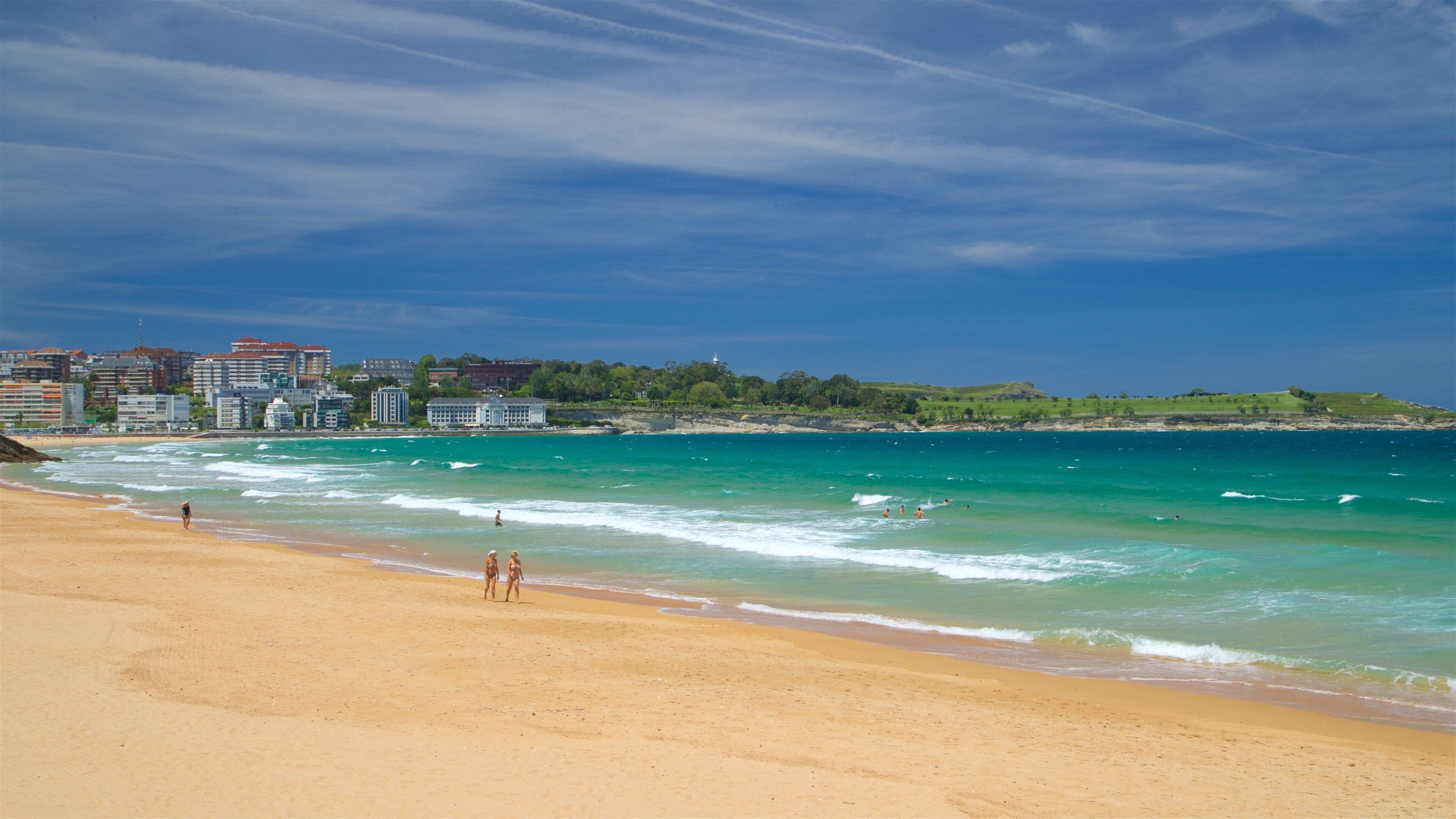 A beach with clear turquoise waters, a sandy shore, and a cityscape in the background.