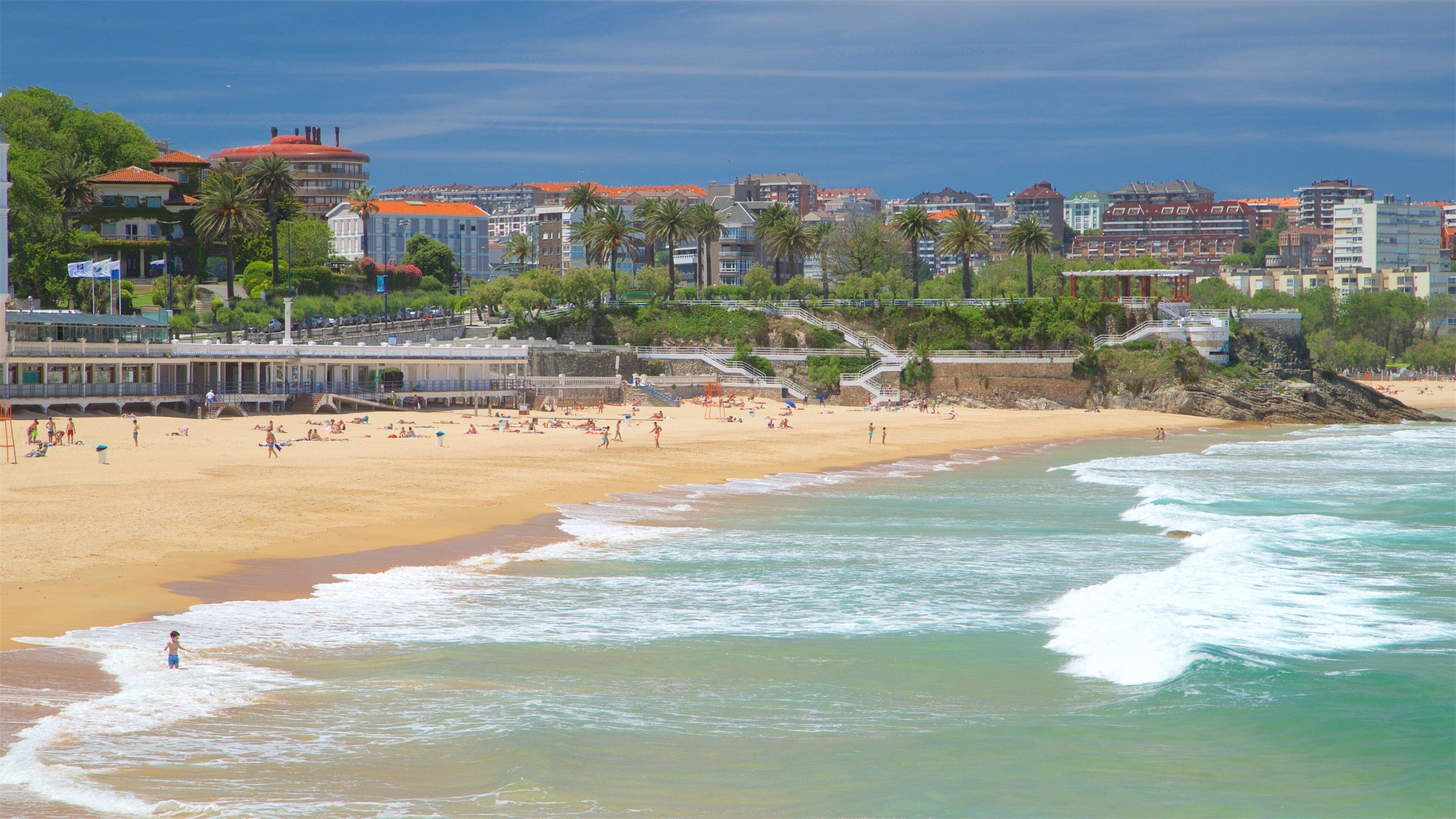 Une plage où des gens se prélassent au soleil et se baignent, une jetée et des bâtiments en arrière-plan.