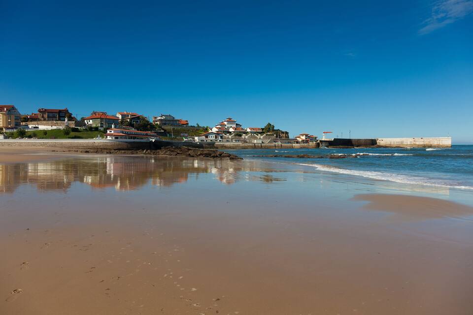 A beach with a calm sea, sandy shore, and houses with red roofs in the background.