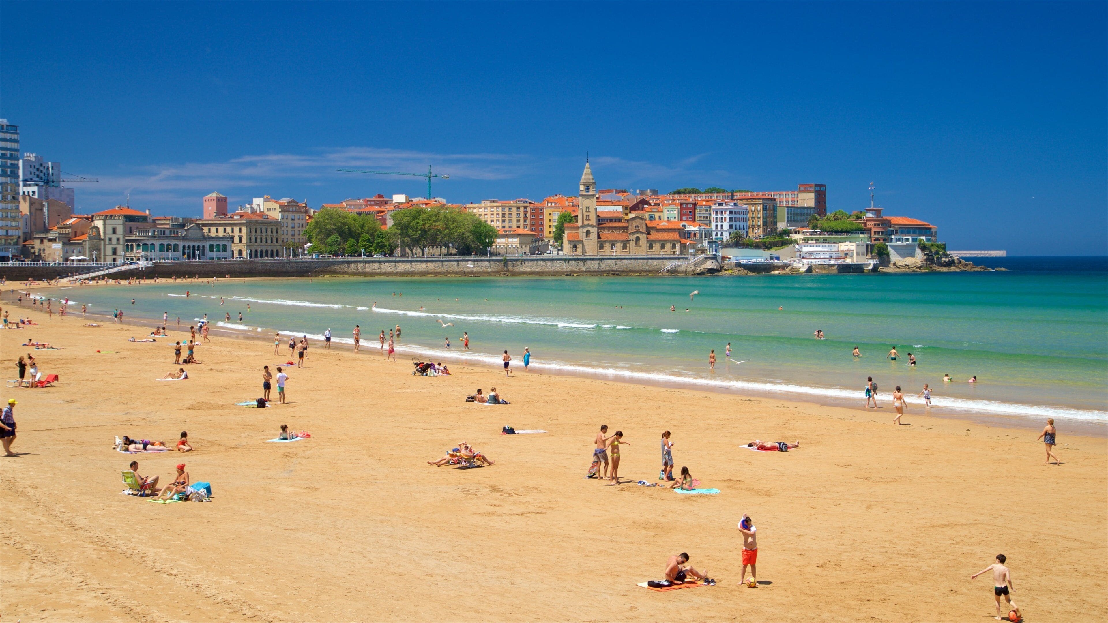 A crowded beach with people sunbathing and playing volleyball. In the background, there are colorful buildings and a tower.