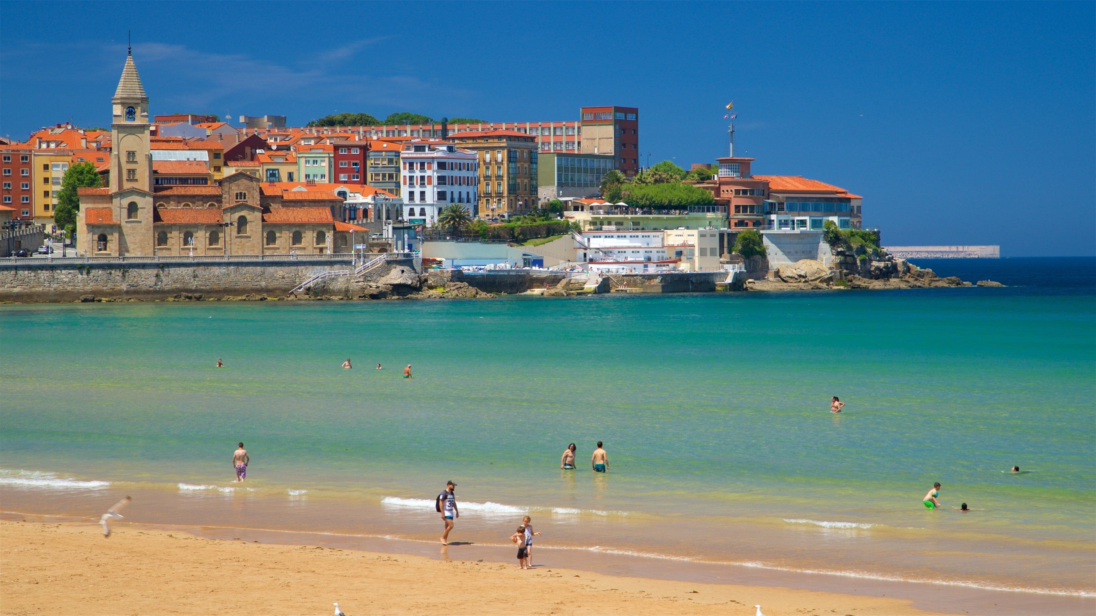 A beach with clear turquoise water, people swimming and walking along the shore, and colorful buildings in the background.
