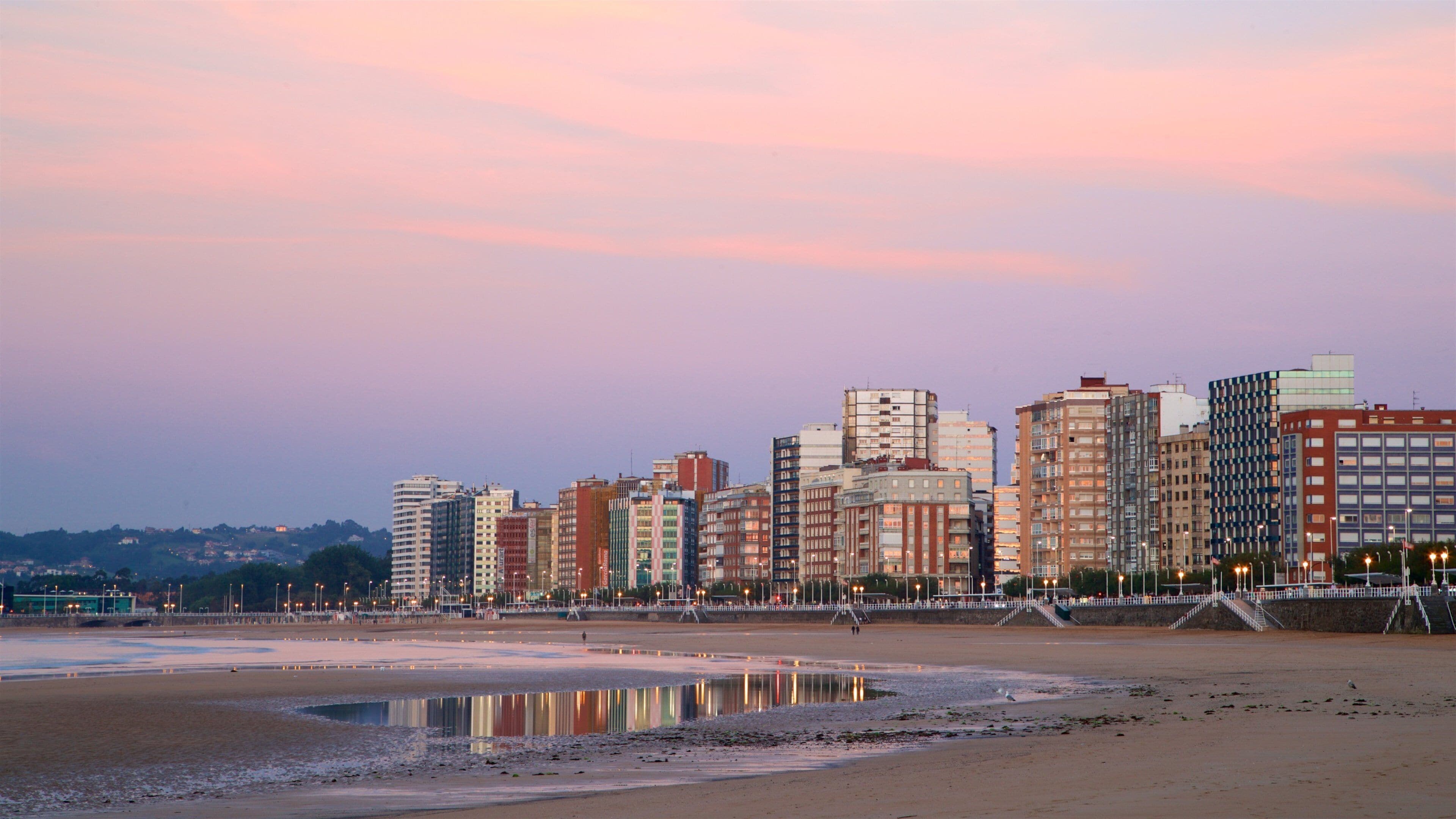 A coastal cityscape with a beach and reflective water at sunset.