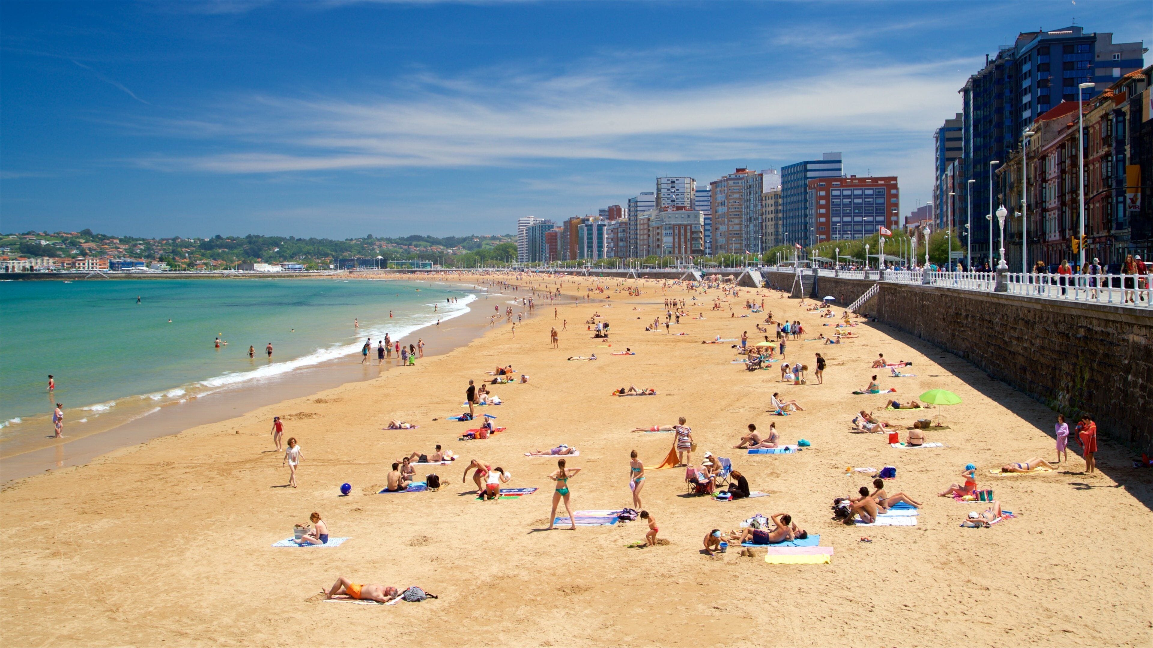 A crowded beach with people sunbathing and swimming, a clear blue sky, and buildings in the background.