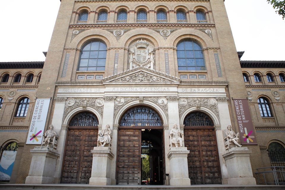 Ein historisches Gebäude mit großen Holztüren und Statuen, mit dem Text 'FACULTADES DE MEDICINA Y CIENCIAS DE ZARAGOZA'.