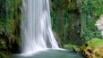 A waterfall cascading down a moss-covered cliff into a calm pool surrounded by lush greenery.