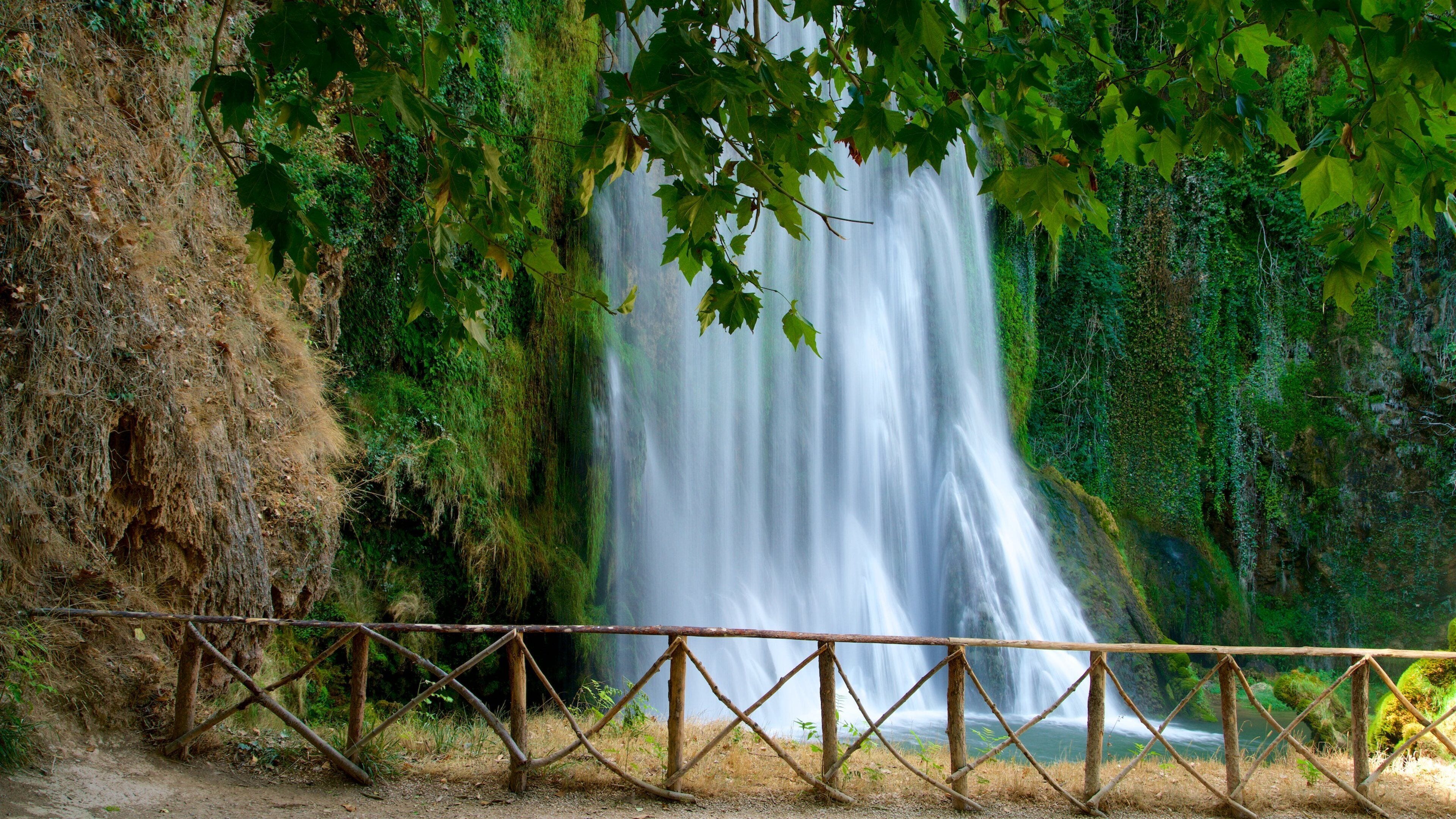 A waterfall surrounded by lush greenery and a wooden fence.