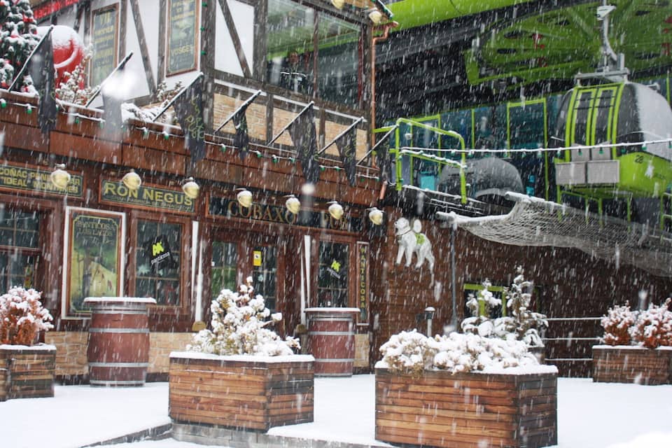 A snowy outdoor scene with a building that has signs in a foreign language, wooden planters with plants, and a green vehicle in the background.