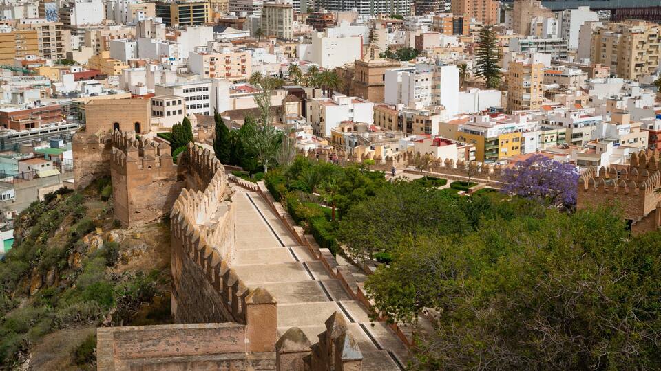 A historic walled city with a prominent fortification, lush greenery, and modern buildings in the background.