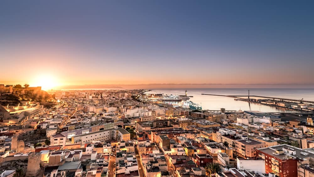 Une vue d’une ville côtière au coucher du soleil, avec un château imposant, un pont et un port de plaisance.