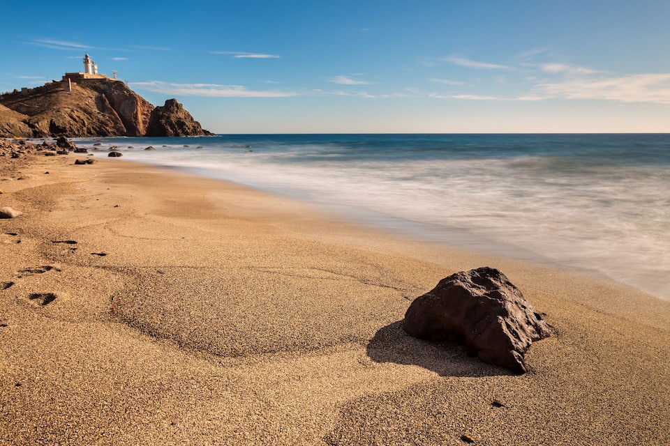 A beach with a large rock in the foreground, a lighthouse on a rocky outcrop, and gentle waves approaching the shore.