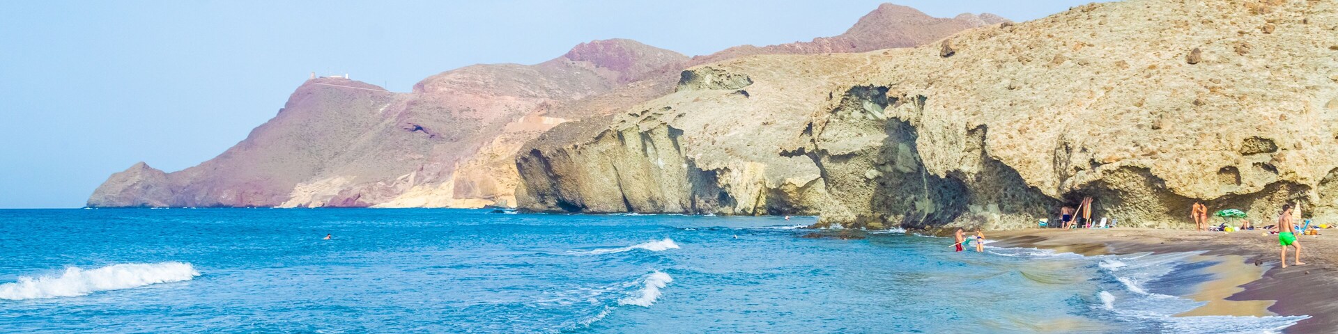 Una playa con aguas azul turquesa, acantilados rocosos y gente disfrutando del paseo marítimo.