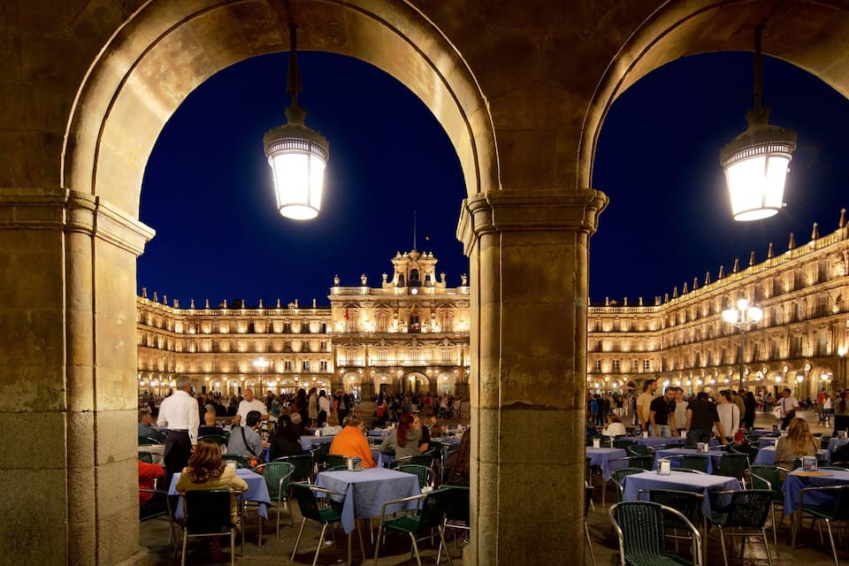 An outdoor café with arches and tables under street lamps, overlooking a historic building with ornate architecture.