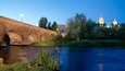 Eine Stebrücke über einem Fluss, mit Bäumen und einem Gebäude mit Kuppeln im Hintergrund.