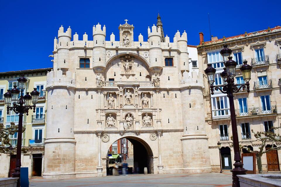 A historic stone gate with intricate carvings and statues, flanked by buildings with balconies and windows.