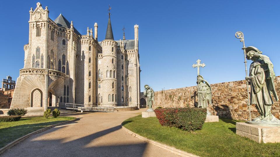 A historic castle with a large central tower, a stone wall, and statues in the foreground.