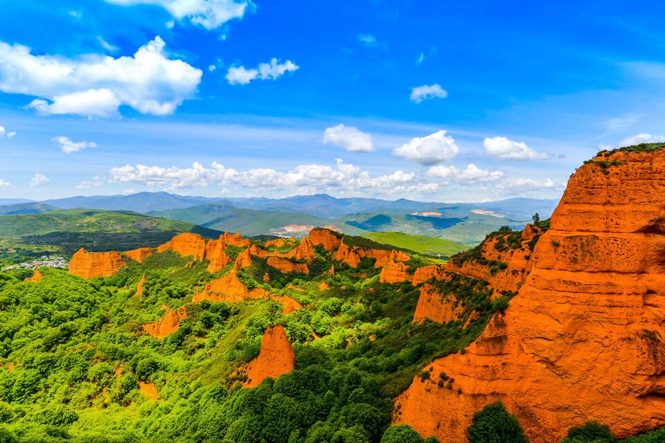 Paesaggio montuoso con formazioni rocciose rosse e vegetazione verde sotto un cielo azzurro.