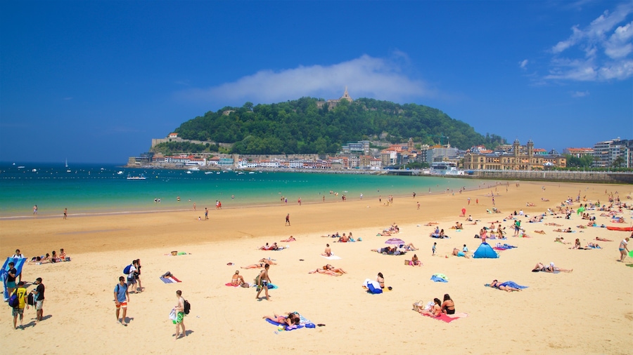 Una playa concurrida con gente tomando el sol y nadando, una colina con edificios al fondo, y un cielo azul despejado.