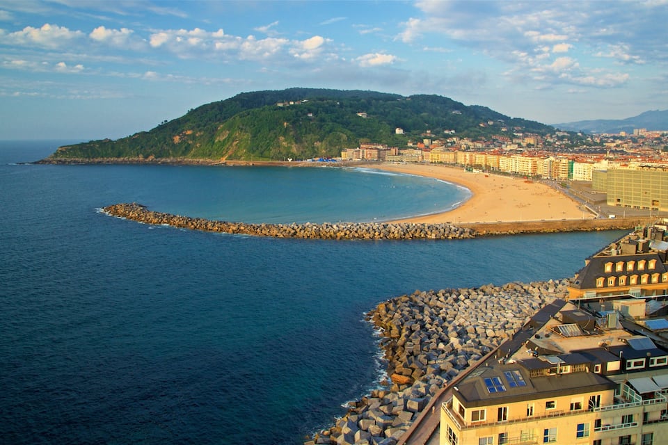 A coastal city with a curved beach, rocky breakwater, and a hill with buildings.