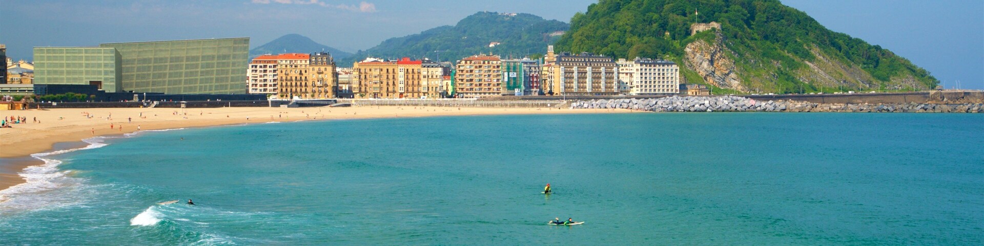 Un paisaje urbano costero con playa, surfistas y una colina con un monumento.