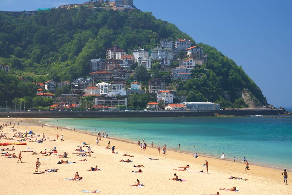 A beach with people sunbathing and swimming, a clear blue sea, and buildings on a hillside.