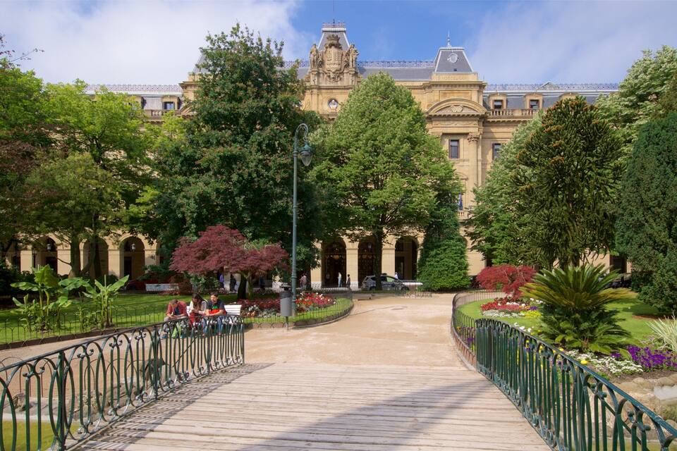 A park with a wooden bridge, lush greenery, and a historic building with ornate architecture.