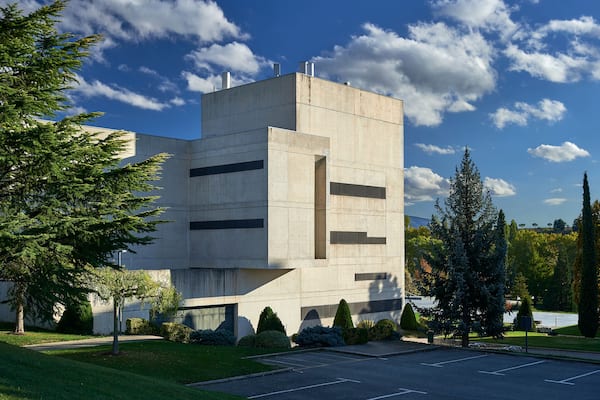 concrete building with horizontal narrow windows, surrounded by pine trees and parking area