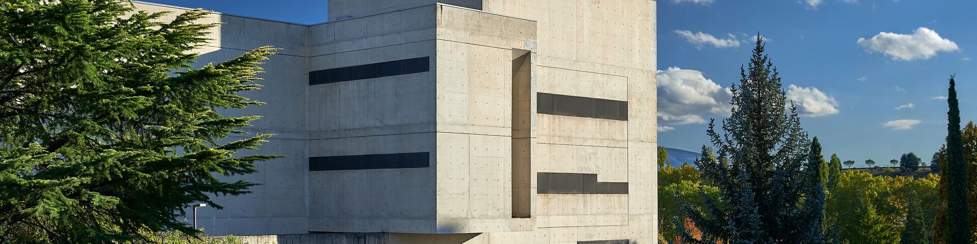 concrete building with horizontal narrow windows, surrounded by pine trees and parking area