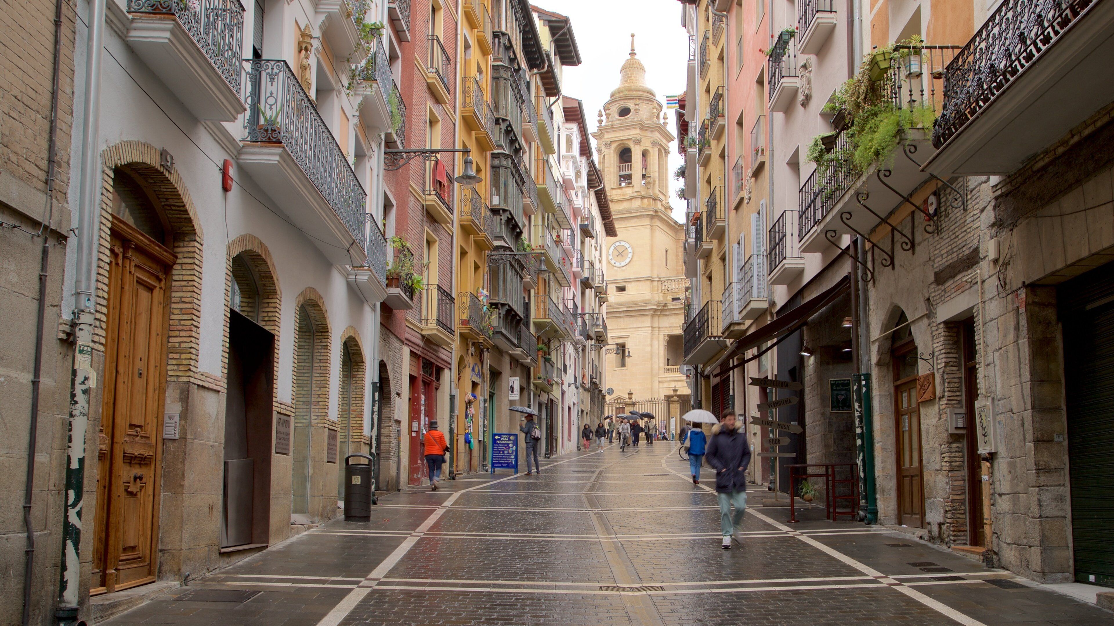 A narrow street lined with colorful buildings, a prominent bell tower, and pedestrians walking.
