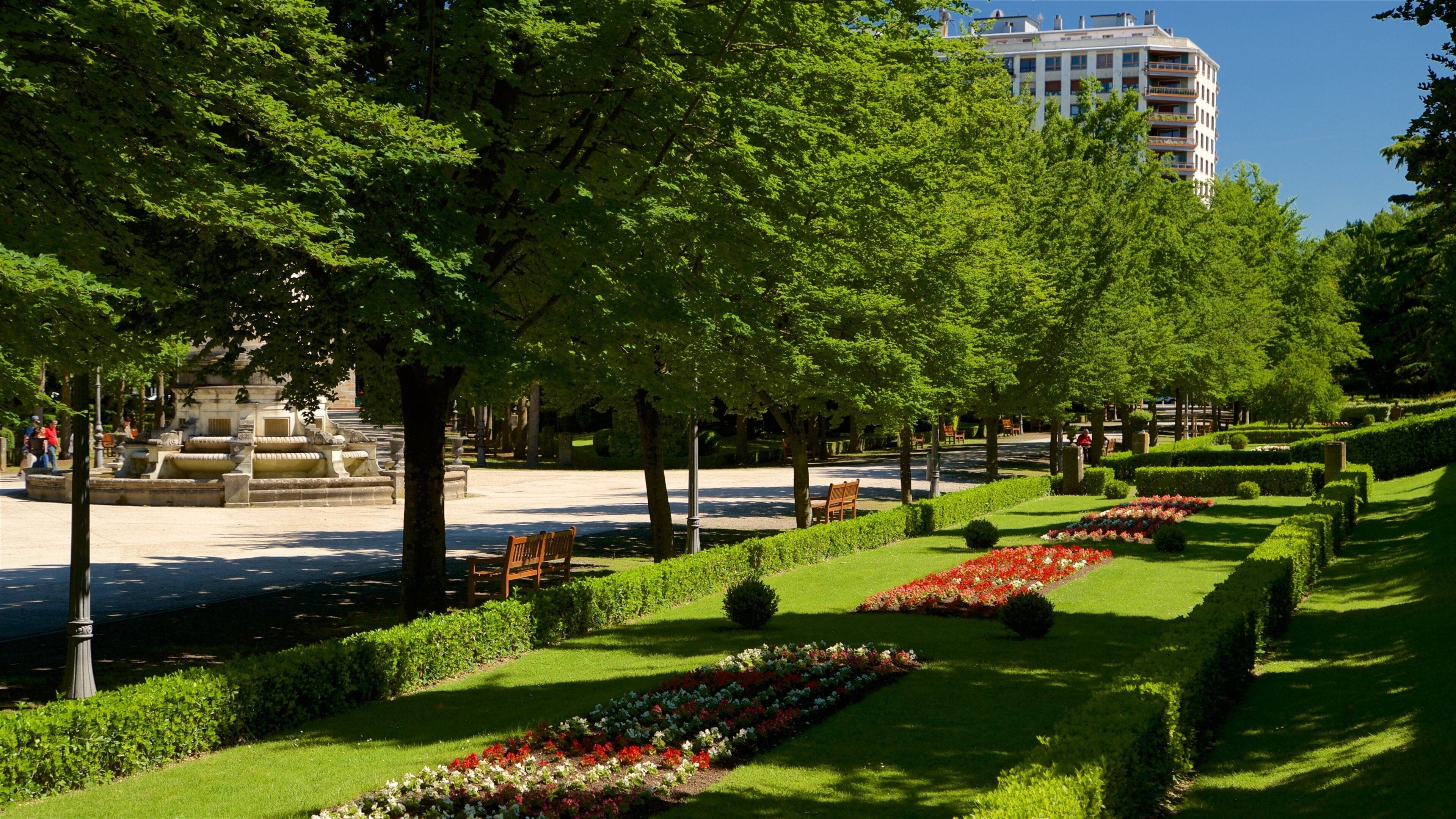 A park with a fountain, neatly trimmed hedges, and a row of colorful flowers.