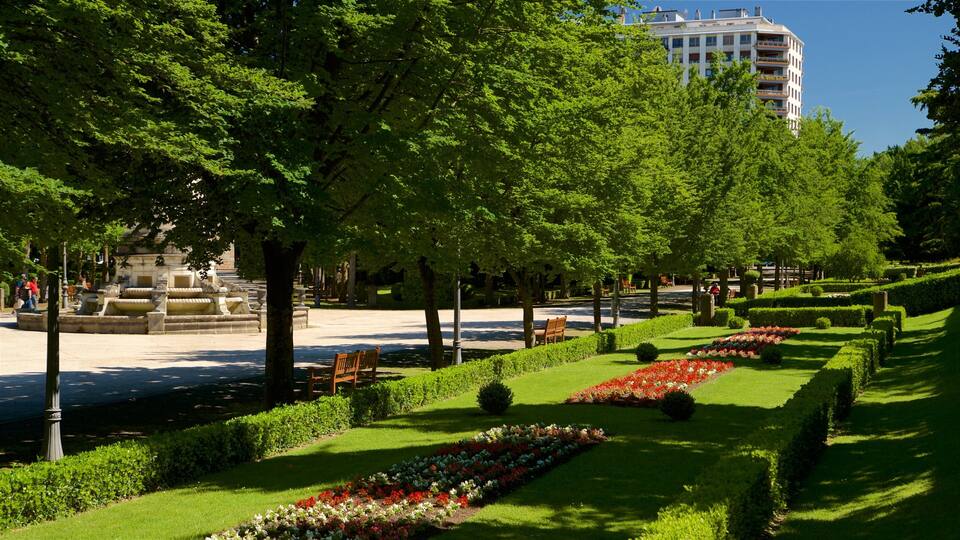 A park with a fountain, neatly trimmed hedges, and a row of colorful flowers.