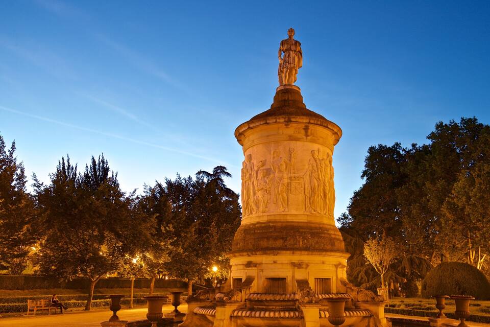 Een goed verlicht monument met een beeld op de top, omringd door bomen en bankjes in een park.