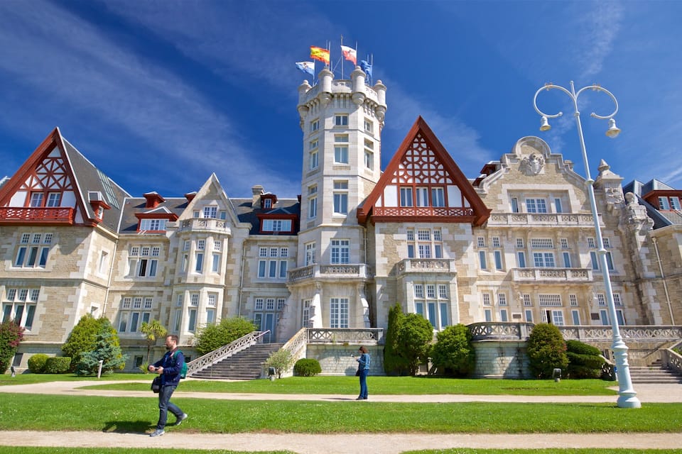 A large, historic building with multiple towers and flags, surrounded by well-maintained lawns and a clear blue sky.