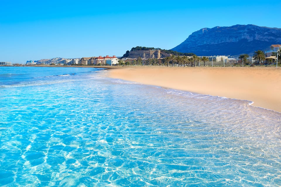 A beach with clear blue water, sandy shore, and a mountain in the background.