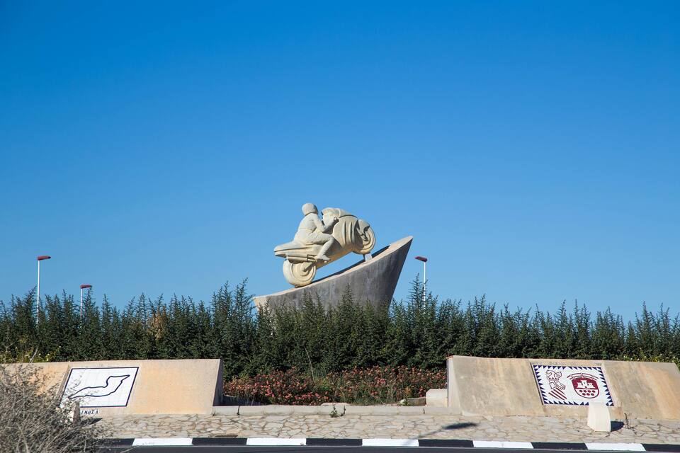 Escultura de uma pessoa a moto sobre uma estrutura ondulada, cercada por árvores, com um emblema de brasão.