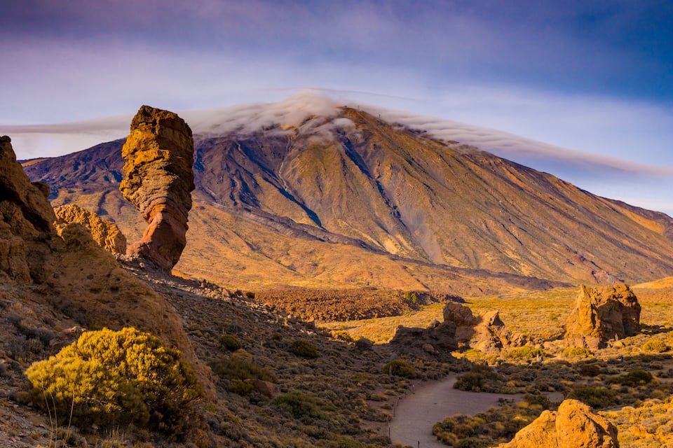 A desert landscape with a prominent rock formation, a mountain in the background, and a trail leading towards the mountain.