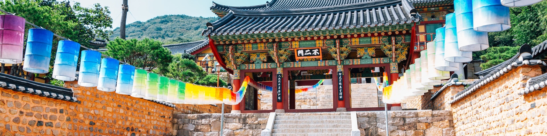 Un portón coreano tradicional con faroles de colores, muros de piedra y montañas al fondo.