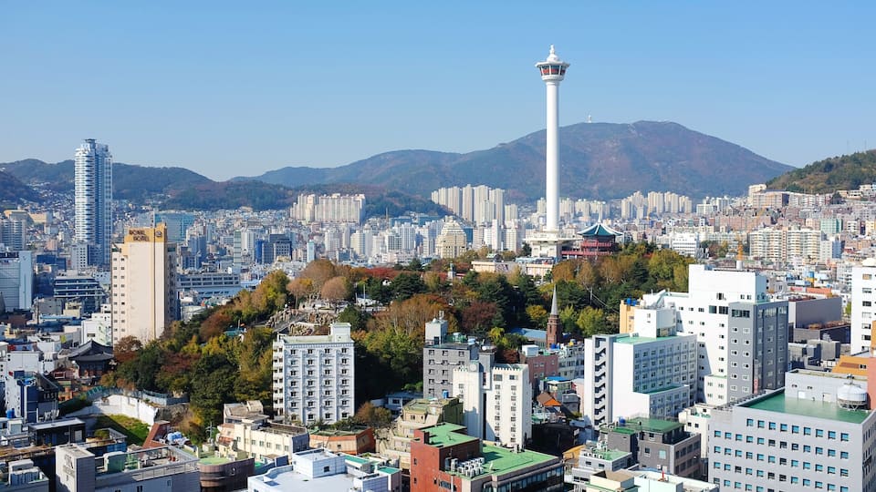 Paisagem urbana com uma torre branca imponente, edifícios urbanos densos e uma montanha ao fundo.